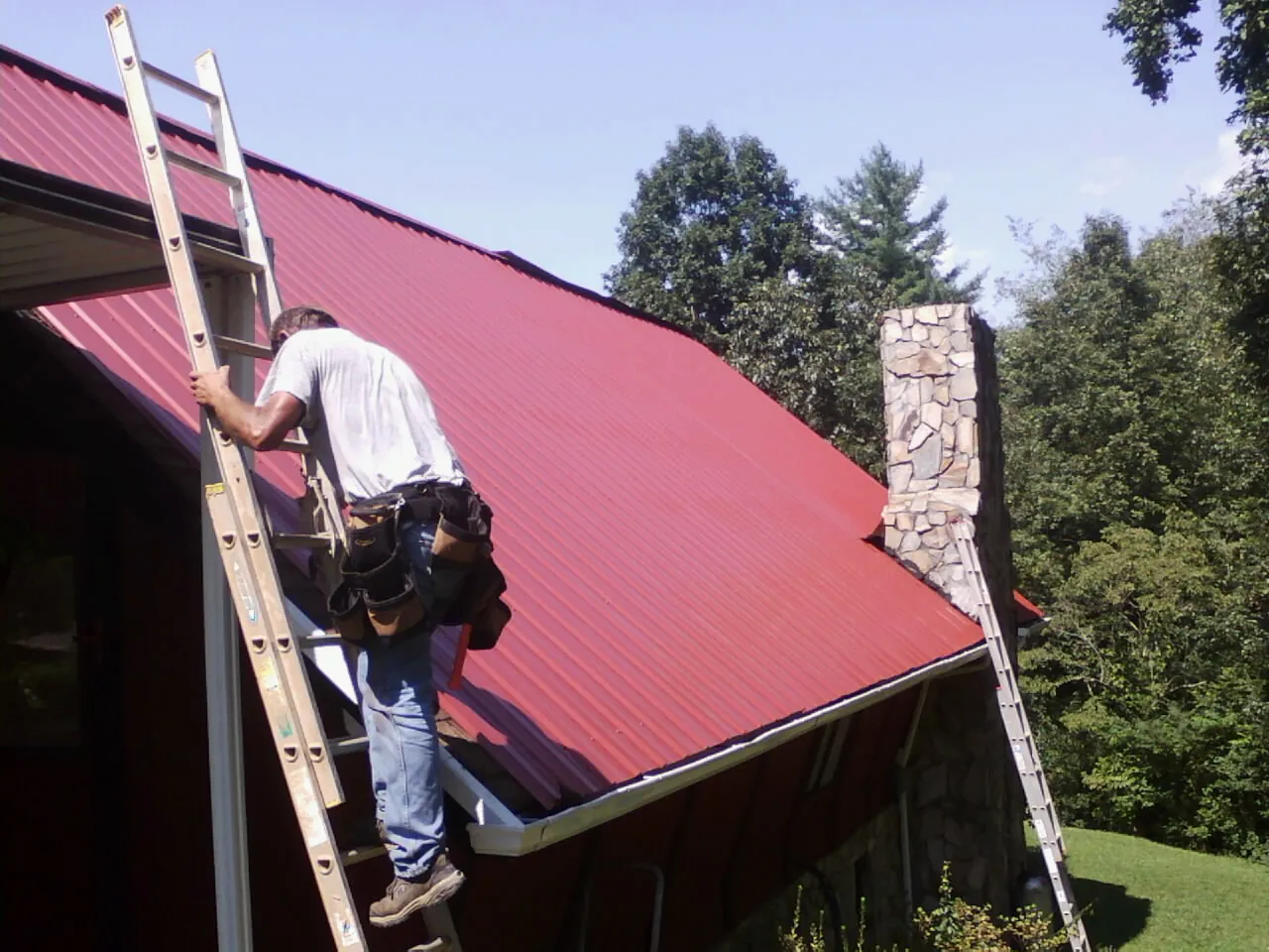 Roofer on a ladder beside a red metal roof and stone chimney — Soffit & Fascia Repair by Roofing Brothers in Fleetville
